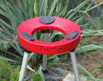 Puede incluir: Un objeto circular rojo con la palabra "ORBIS" grabada, sostenido por tres patas plateadas. El objeto tiene tres almohadillas negras y circulares en su superficie superior. La imagen está tomada al aire libre, con vegetación de fondo.