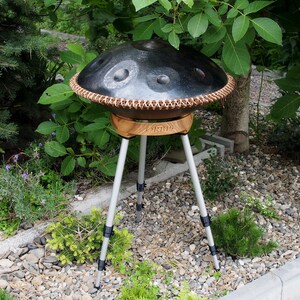 May include: A black handpan drum with a copper-colored rim and a wooden base, supported by a silver tripod stand. The instrument is outdoors, surrounded by greenery and small rocks.