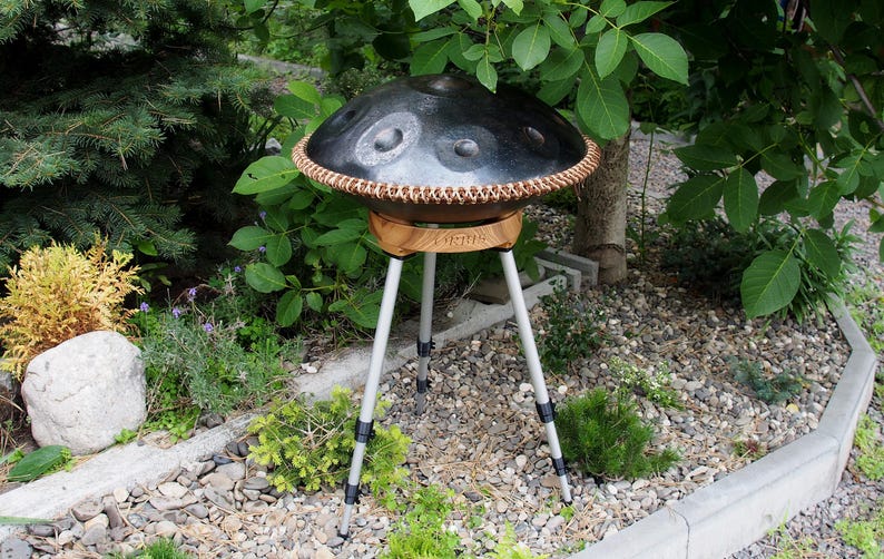 May include: A black handpan drum with a copper-colored rim and a wooden base, supported by a silver tripod stand. The instrument is outdoors, surrounded by greenery and small rocks.