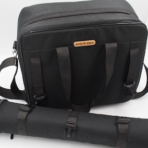 May include: A black padded carrying case with a shoulder strap and a separate cylindrical case. The carrying case has a zipper closure and a brown label with the word "ORTO" printed on it.