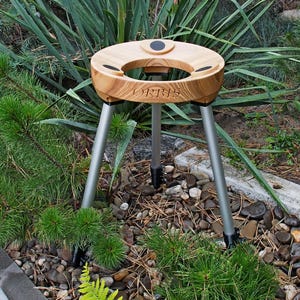 May include: A wooden outdoor table with a round top and three silver legs. The table has a cutout in the center and is surrounded by green plants and small rocks. The table has the word "URBIS" engraved on it.