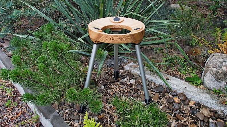 May include: A wooden outdoor table with a round top and three silver legs. The table has a cutout in the center and is surrounded by green plants and small rocks. The table has the word "URBIS" engraved on it.