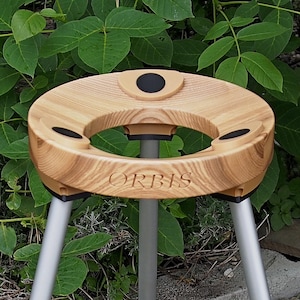 May include: A wooden tripod with a circular top, featuring the engraved word "ORBIS". The top has a central opening and three black circular insets. The tripod legs are silver-colored and the background is lush green foliage.
