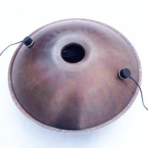 Może przedstawiać: Brązowy stalowy handpan z centralnym otworem dźwiękowym i dwoma czarnymi przetwornikami przymocowanymi przewodami. Instrument ma okrągły kształt i teksturowaną powierzchnię, na białym tle.