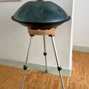May include: A teal handpan drum with a black rim, resting on a wooden stand with the word "ORBIS" engraved on it. The stand has three silver legs. The instrument is in a room with a wooden floor.
