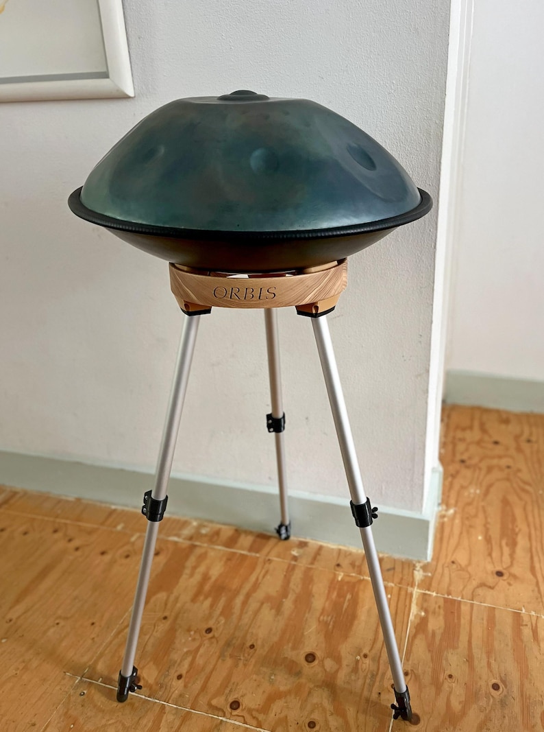May include: A teal handpan drum with a black rim, resting on a wooden stand with the word "ORBIS" engraved on it. The stand has three silver legs. The instrument is in a room with a wooden floor.