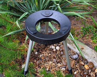 Puede incluir: Un soporte de planta trípode negro con una parte superior circular y tres patas plateadas. El soporte tiene tres pequeñas almohadillas negras redondas en la parte superior. El soporte está en un jardín con plantas verdes y pequeñas rocas.