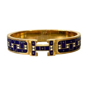 May include: A gold-tone hinged bangle bracelet featuring a central H-shaped clasp. The exterior surface is decorated with a dark blue enamel inlay, accented by a repeating white and grey geometric pattern that wraps around the entire band.