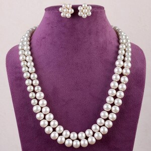 Peut inclure: Un collier de perles à deux rangs avec une paire de boucles d'oreilles assorties. Le collier et les boucles d'oreilles sont blancs et sont présentés sur un support en velours violet.