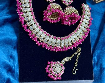Puede incluir: Un conjunto de joyas de novia indias de color rosa y blanco con un collar, pendientes y un aro para la nariz. El collar está hecho de oro y tiene un diseño floral con piedras rosas y blancas. Los pendientes también están hechos de oro y tienen un diseño similar. El aro para la nariz es un simple aro de oro con una piedra rosa.