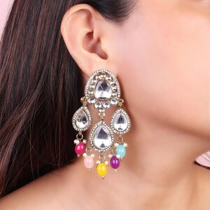 May include: Gold tone dangle earrings with clear crystal stones and colorful beads. The earrings feature a teardrop shape with a large crystal stone in the center and smaller stones surrounding it. The beads are pink, yellow, blue, and purple.
