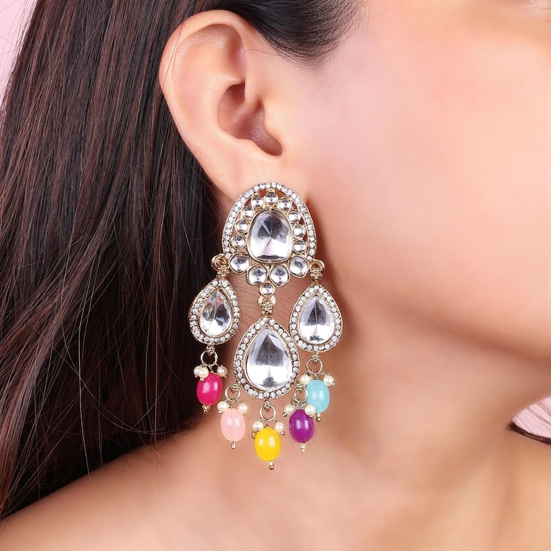 May include: Gold tone dangle earrings with clear crystal stones and colorful beads. The earrings feature a teardrop shape with a large crystal stone in the center and smaller stones surrounding it. The beads are pink, yellow, blue, and purple.