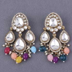 May include: A pair of gold-tone earrings with a teardrop design. The earrings feature clear stones and colorful beads in shades of pink, blue, yellow, and purple.
