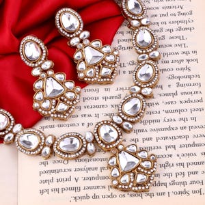 May include: Gold-toned jewelry set featuring a necklace and earrings. The pieces are adorned with clear, oval and triangular-shaped stones, set in a detailed, ornate design. The jewelry is displayed on a red fabric and a book.