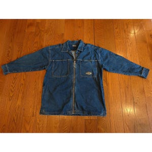 Könnte beinhalten: Blaue Jeansjacke mit durchgehendem Reißverschluss. Die Jacke hat zwei Brusttaschen und einen kleinen Logoaufnäher auf der rechten Tasche. Die langärmlige Jacke liegt auf einer Holzoberfläche.