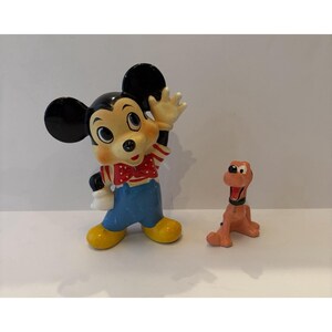 May include: A ceramic figurine of a cartoon mouse with large black ears, wearing a red and white polka-dot bow tie, a striped shirt, and blue overalls, standing next to a smaller, light brown cartoon dog figurine with a black collar.