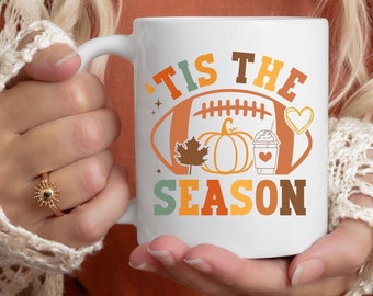 May include: White ceramic mug with the text "'Tis the Season" in brown and orange lettering. A football with a pumpkin, a leaf, and a coffee cup are inside the football.