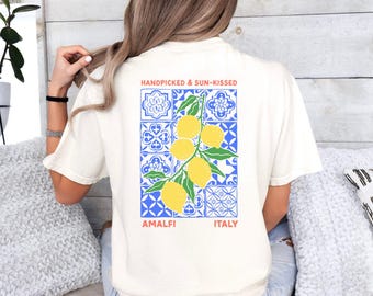 May include: Cream-colored t-shirt featuring a graphic of lemons on a branch, set against a blue and white tile pattern. The text "HANDPICKED & SUN-KISSED" and "AMALFI ITALY" are printed above and below the graphic.