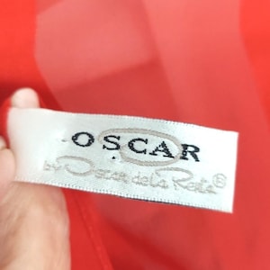 May include: A close-up of a white label with the text "OSCAR" in black, inside a gray oval. Below the text is "by Oscar de la Renta" in a cursive font. The label is against a red background.