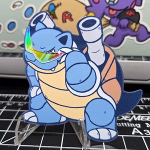 May include: A blue and yellow cartoon sticker of a turtle-like creature with a rainbow visor. The sticker is on a clear stand, set against a black cutting mat with white grid lines. Other stickers are visible in the background.