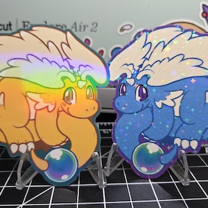 Puede incluir: Dos pegatinas holográficas con dibujos de dragones. Una es naranja con alas de arcoíris, la otra es azul con purpurina. Cada dragón tiene una burbuja y está sobre un soporte transparente. Las pegatinas están sobre una superficie negra.