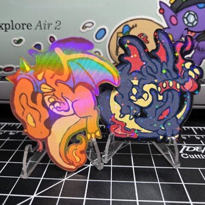 May include: Two holographic stickers of cartoon dragons, one orange and yellow, the other dark blue and red, are displayed on clear stands. A purple cartoon character with a hat is visible in the background. The stickers are on a black cutting mat.