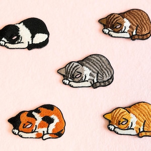 May include: Embroidered patches of sleeping cats in various colours, including black and white, brown and white, grey, calico, and orange and white. The patches are on a light pink background.