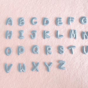 May include: A full set of light blue embroidered alphabet letters, from A to Z, displayed on a soft pink surface. Each letter has a puffy, textured appearance, creating a three-dimensional effect. The letters are evenly spaced.