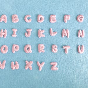 Può includere: Lettere dell'alfabeto in tessuto rosa disposte su uno sfondo di feltro azzurro. Le lettere sono in un carattere arrotondato, in stile bolla. Le lettere sono disposte in righe, che compongono l'alfabeto.