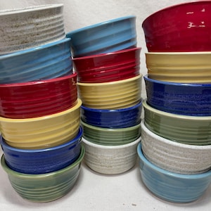 Può includere: Una pila di 12 ciotole in ceramica di vari colori, tra cui blu, rosso, giallo, verde e bianco. Le ciotole hanno un design testurizzato e nervato.