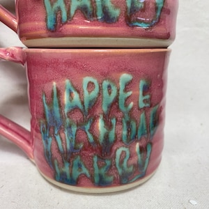 May include: Two pink ceramic mugs with a teal glaze. The top mug says "Happy Birthday Harley" and the bottom mug says "Happy Birthday Harley".
