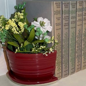 May include: A red ceramic flower pot with a matching saucer holds an arrangement of artificial flowers. The flowers are white, yellow, and green. To the right are a series of vintage Nancy Drew Mystery Stories books with decorative spines.
