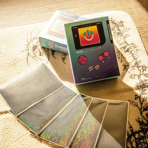 以下が含まれることがあります： ホログラフィックカードスリーブのセットと「Classic Cards」の2つの箱。箱は、電源ボタンのグラフィックが付いたレトロな携帯ゲーム機のようにデザインされています。箱はティール、紫、ピンクで、「Retro Fila」の文字が入っています。