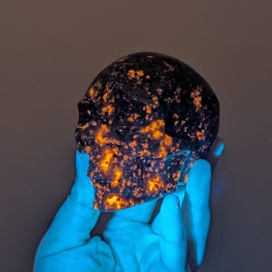 May include: A carved skull made of a dark, speckled stone with orange and yellow inclusions. The skull is held in a gloved hand, and the overall effect is a glowing, otherworldly appearance. The skull is a decorative item.