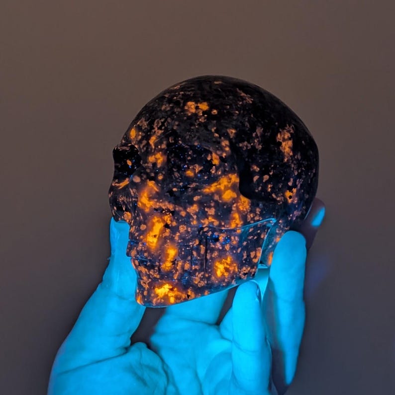 May include: A carved skull made of a dark, speckled stone with orange and yellow inclusions. The skull is held in a gloved hand, and the overall effect is a glowing, otherworldly appearance. The skull is a decorative item.