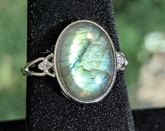 Puede incluir: Anillo de plata con una piedra de labradorita ovalada e iridiscente, que muestra tonos verdes y azules. El anillo presenta pequeñas piedras de acento transparentes y un diseño de nudo celta en la banda.