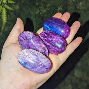 May include: Three polished, oval-shaped stones with a smooth, reflective surface. The stones display vibrant shades of deep purple, magenta, and violet, with iridescent blue flashes visible on the surface of the stones, showcasing natural mineral textures.