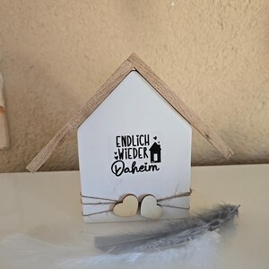 Puede incluir: Un adorno de madera blanco en forma de casa con el texto "ENDLICH WIEDER DAHEIM" en letras negras. Dos pequeños corazones de madera están unidos a la parte inferior de la casa con un cordel. Una pluma blanca está sobre una superficie blanca en primer plano.