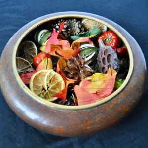 May include: A round, brown, textured bowl filled with autumn-themed potpourri, including dried orange slices, pinecones, red artificial berries, green leaves, and orange and red faux maple leaves, all arranged inside the container.