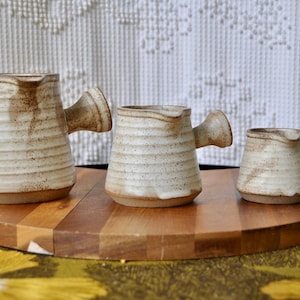 May include: Three handmade ceramic pitchers in varying sizes, arranged on a wooden serving board. The pitchers are beige with brown speckles and horizontal ridged detailing. Each has a small, curved handle and a spout.