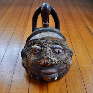 Puede incluir: M&aacute;scara tribal artesanal de color marr&oacute;n oscuro, con una superficie texturizada y detalles intrincados. La m&aacute;scara presenta un rostro estilizado con ojos abiertos, una boca peque&ntilde;a y un patr&oacute;n decorativo en la frente. Un lazo se extiende desde la parte superior.