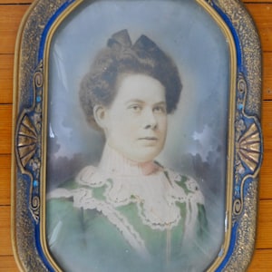 May include: Oval portrait of a person in a green dress with white lace trim, set within a decorative frame. The frame has a gold and blue design with ornate detailing. The portrait is behind glass.