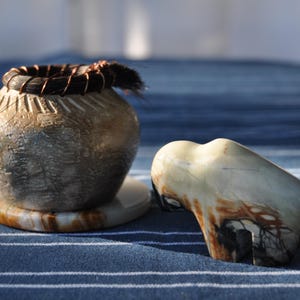 May include: A small, round, textured gray pot with a dark rim wrapped in copper wire, resting on a circular stone base, accompanied by a smooth, carved stone figurine of a buffalo featuring cream, brown, and black marbled patterns.