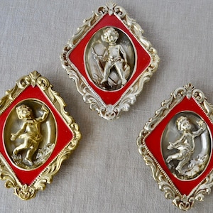 May include: Three vintage wall plaques featuring cherub figures. Each plaque has a red center with a gold-colored cherub relief, set within a decorative gold and silver-colored frame. The plaques are diamond-shaped.