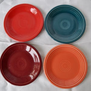 May include: Four colorful ceramic plates in red, teal, burgundy, and orange. Each plate has a concentric ring design in the center and a slightly raised rim. The plates are arranged on a white surface, showcasing their vibrant hues.