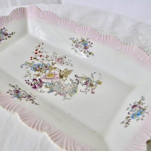 May include: Rectangular white ceramic serving dish with a scalloped pink rim and floral design. The dish features a central bouquet of flowers in shades of pink, blue, and yellow, with smaller floral clusters around the edges.