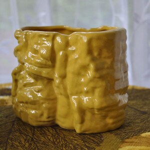 May include: A mustard-yellow ceramic vase with a textured, rough surface. The vase has a rectangular shape with rounded corners and a wide opening. The design appears to be inspired by a tree trunk or bark.