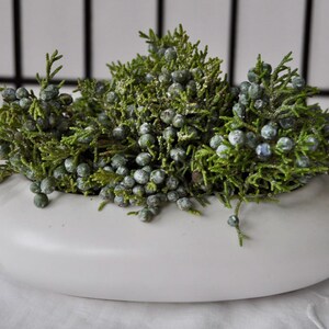 May include: A low, wide, white ceramic bowl filled with dense, green juniper branches covered in small, round, dusty blue-green berries. The arrangement sits compactly within the smooth, rounded basin, creating a contrast between the organic texture of the foliage and the clean, minimalist white surface of the container.