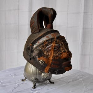 Puede incluir: M&aacute;scara de madera antigua con rostro tallado, de color marr&oacute;n oscuro con detalles rojos y blancos. La m&aacute;scara est&aacute; montada sobre una base de metal plateado con tres patas decorativas. La m&aacute;scara tiene una apariencia &uacute;nica y envejecida.