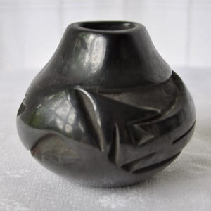 May include: A small, polished black pottery vessel with a rounded body and a narrow opening. The surface features carved, angular geometric indentations that create a textured pattern around the midsection of the dark, glossy ceramic object.